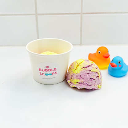 Bath bomb with a container labeled 'Bubble Scoops' and two rubber duck toys on a tiled bathroom floor.