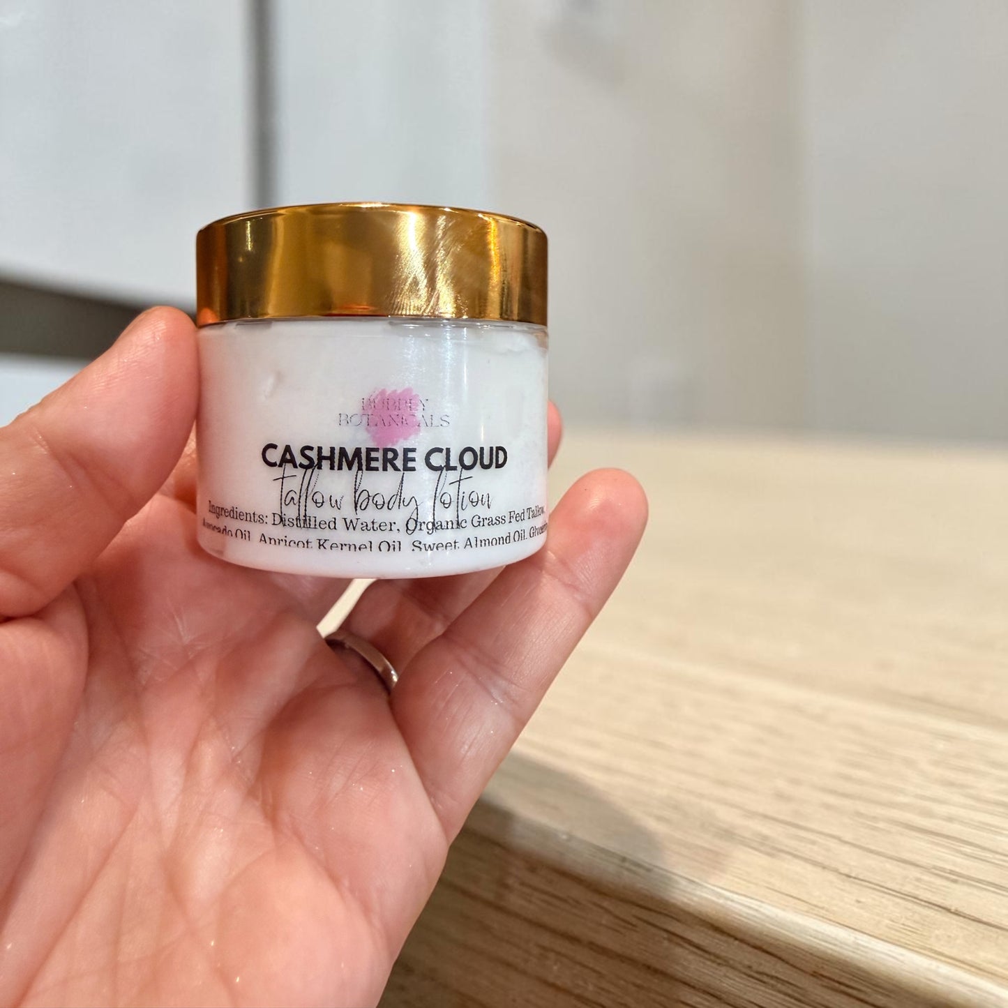 A hand holding a jar of Cashmere Cloud Tallow Body lotion against a blurred indoor background.