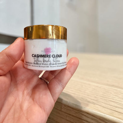 A hand holding a jar of Cashmere Cloud Tallow Body lotion against a blurred indoor background.