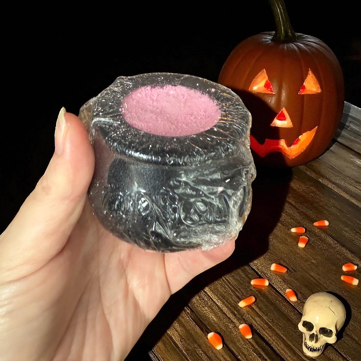Hand holding a black Halloween-themed cauldron bath bomb with pink center, jack-o'-lantern, and skull in the background.