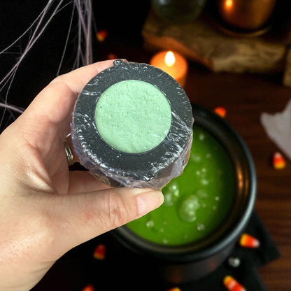 Hand holding a wrapped green cauldron bath bomb with a Halloween-themed background