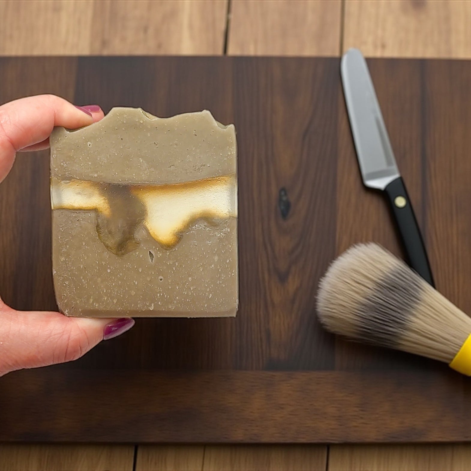 Hand holding a bar of soap with a knife and brush on a wooden surface