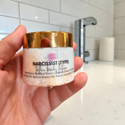 A hand holding a small jar of tallow body lotion labeled 'NARCISSIST (TYPE) Tallow BODY Lotion against a blurred bathroom background with a sink and tiled wall.