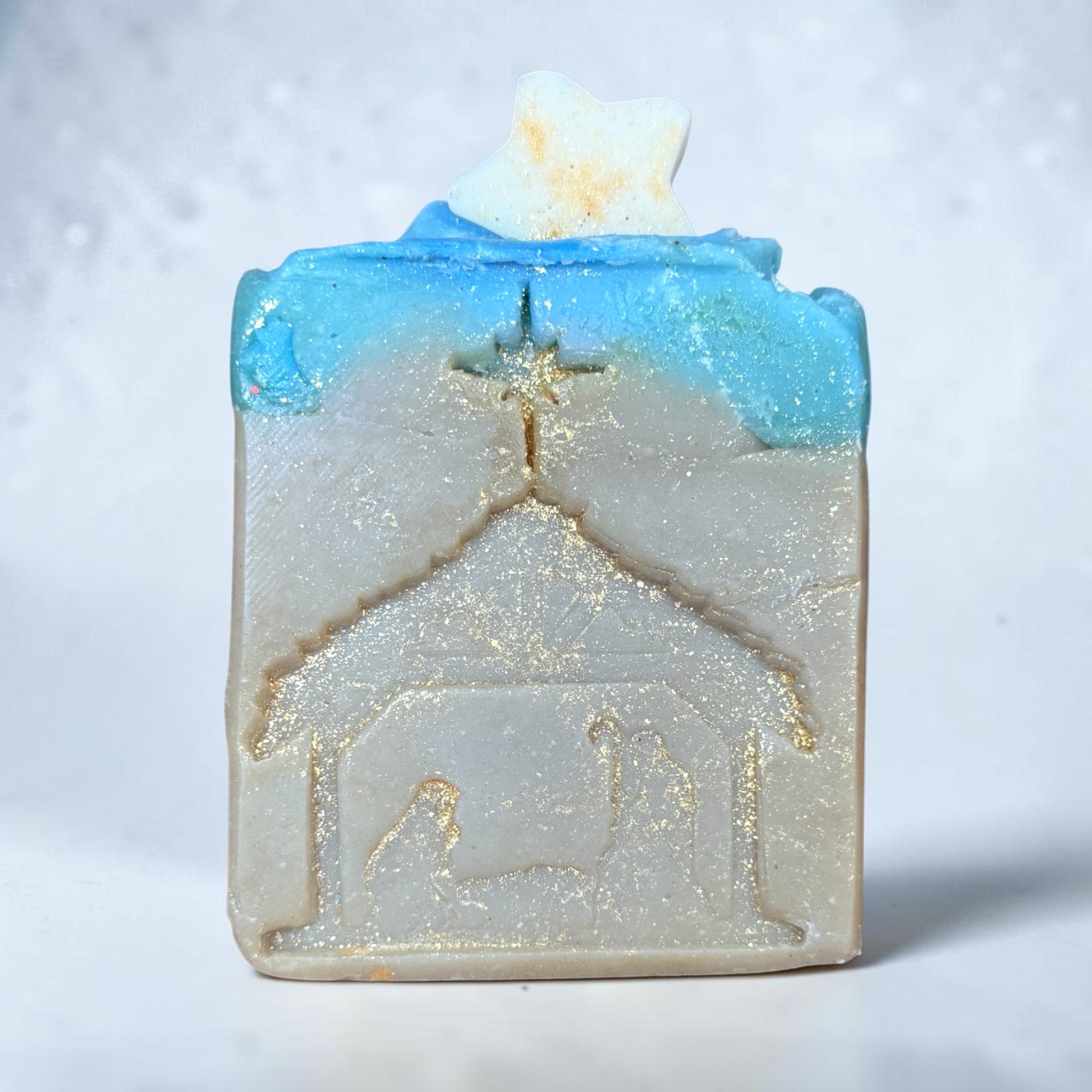 A small rectangular artisan tallow soap with a nativity scene embossed on the front, featuring a blue and white gradient top with a gold glitter finish, set against a light, sparkly background.