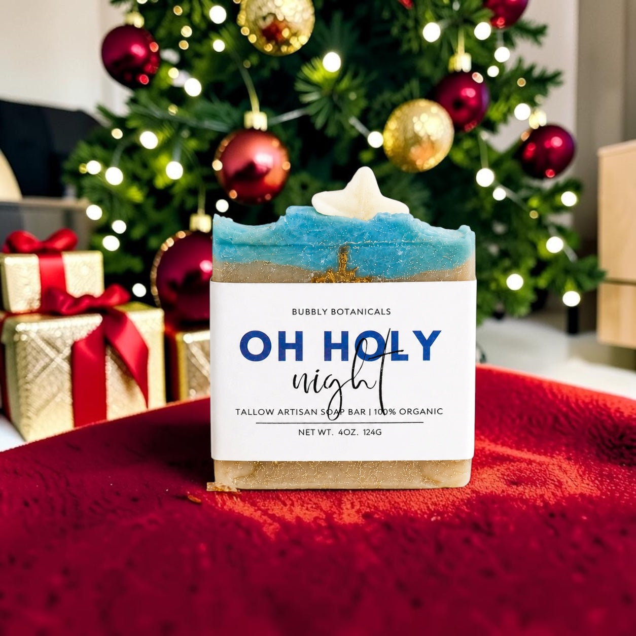 A handcrafted bar of tallow soap labeled 'OH HOLY night' by Bubbly Botanicals, featuring a blue and beige marbled top with a white star topper, set on a red textured surface with a decorated Christmas tree and gifts in the background.