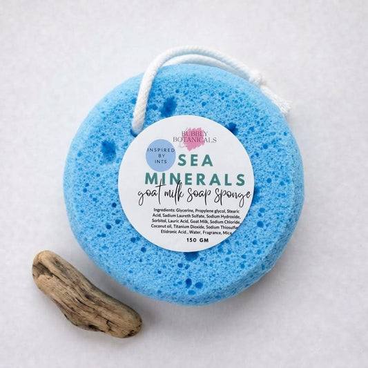 Sea Minerals goat milk soap infused sponge by Bubbly Botanicals, a round blue sponge with label beside a piece of driftwood
