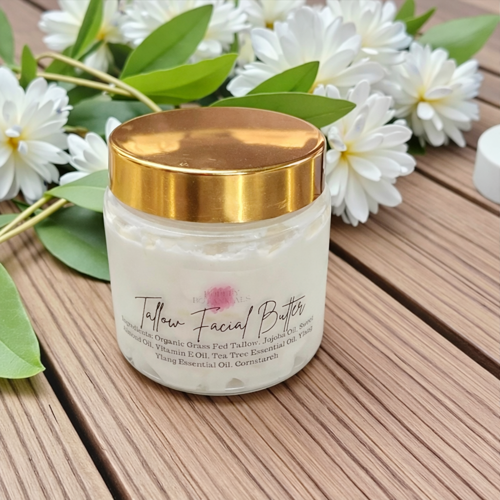 Jar of Tallow Facial Butter with a gold lid on a wooden surface with white flowers and green leaves.