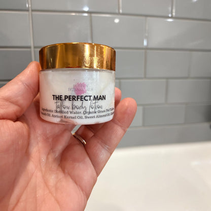 A hand holding a small white cosmetic jar with a metallic gold lid. The label reads 'THE PERFECT MAN' tallow body lotion mini